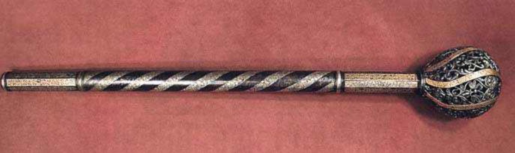 Hetman Pylyp Orlyk's mace, 18th century. A long silver and black staff with an intricately carved wooden pommel on the end, decorated in gold filigree, lying horizontally against a red background. The stick has five straight horizontal stripes running along its length, each striped in different patterns or colors, creating a sense of depth and texture. A small ball-shaped decoration is attached to one side at about half way from view.